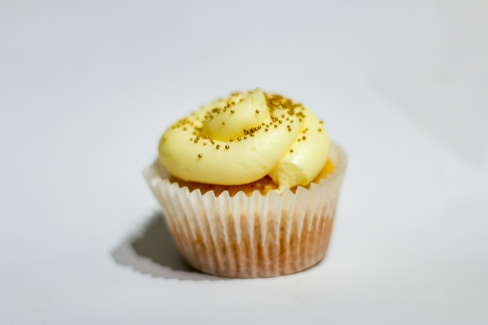A cupcake with yellow frosting and small gold sprinkles in a white paper case
